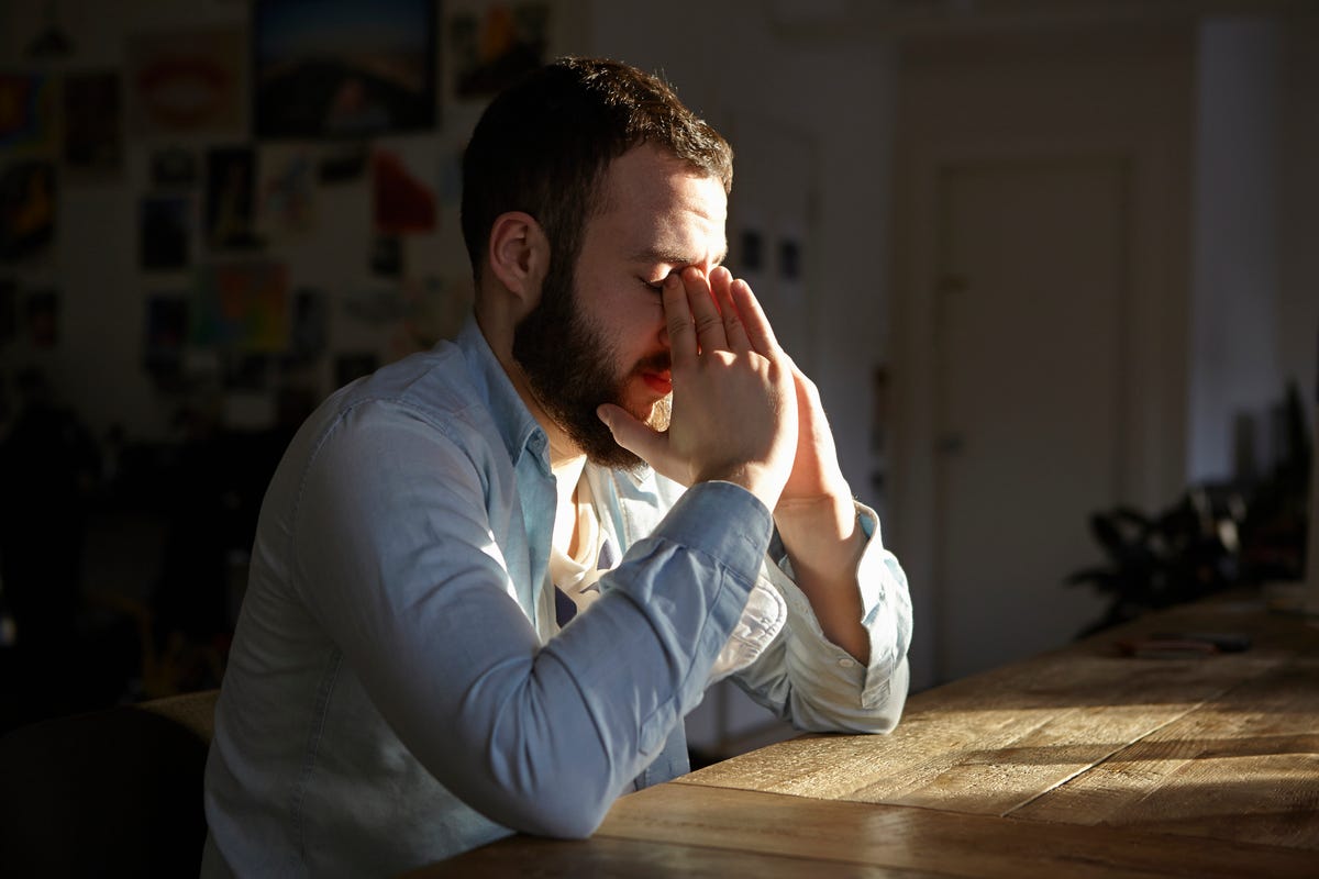 Man sitting at kitchen table with hands on face showing distress.