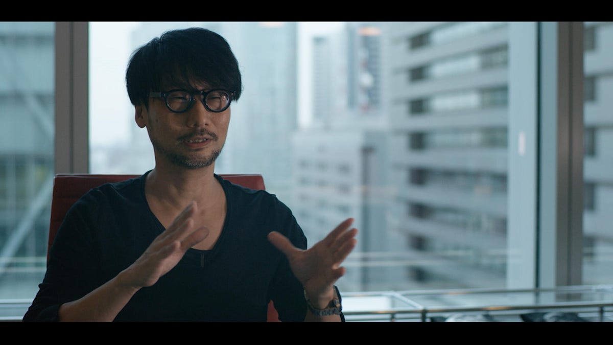 Hideo Kojima, sitting in a chair, gestures with his hands