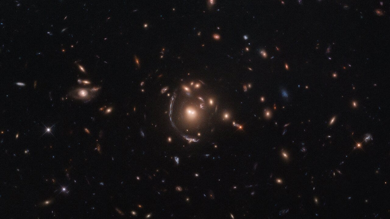 A dark background is scattered with hazy, glowing galaxies. At the center, one galaxy appears stretched out as a long arc instead of having a characteristic spiral, for instance.