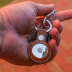 A hand holds keys with an Apple AirTag attached via a leather loop keyring.