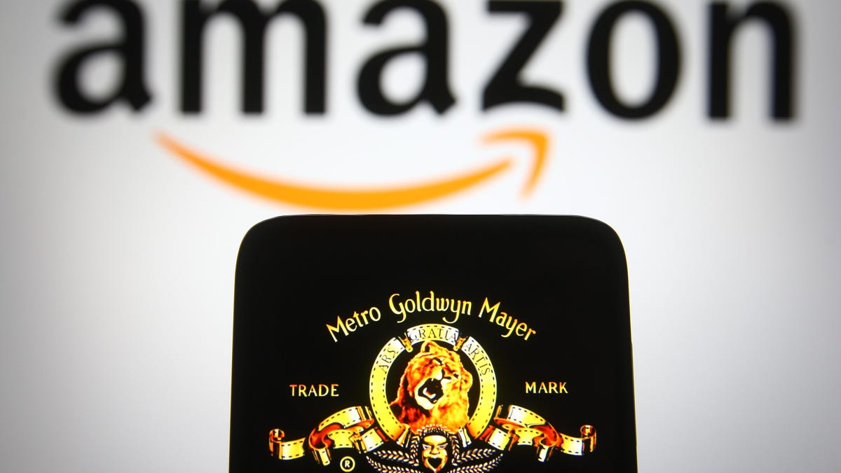 A phone displays the MGM lion logo. Behind the phone is a larger, blurred out Amazon sign.