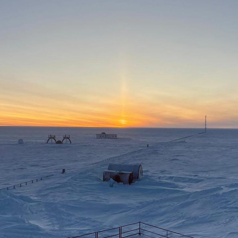Snowy expanse of Antarctica with fencing and outbuildings. The sun ends up a streak of light at the horizon.