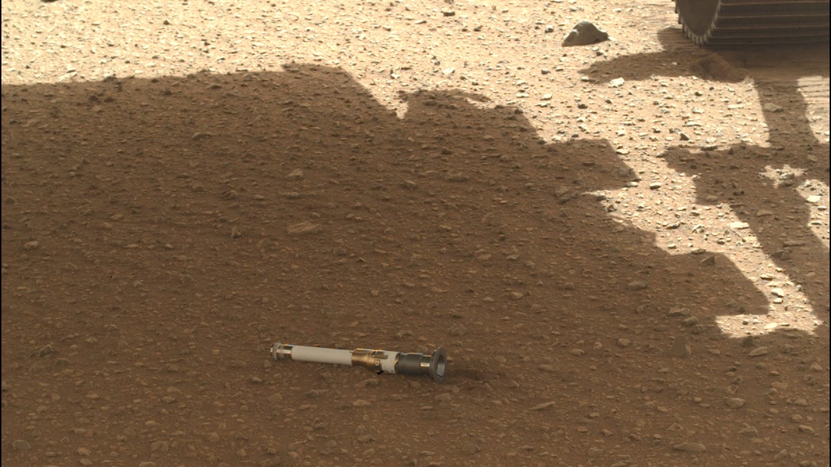 a white tube lies on the surface of Mars, in the shadow of the Perseverance rover. A wheel is partially visible in the top right hand corner.