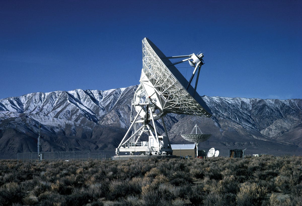 In the background, there's a medium blue sky, a mountain range that runs perpendicular to the photo's landscape and bushes in the foreground. Right in the center of this scene is a giant white instrument that holds one of the VLBA's antennas.