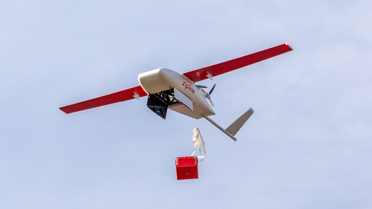 A Zipline delivery drone, looking like a small airplane, drops a red box with a plastic parachute attached to slow its fall