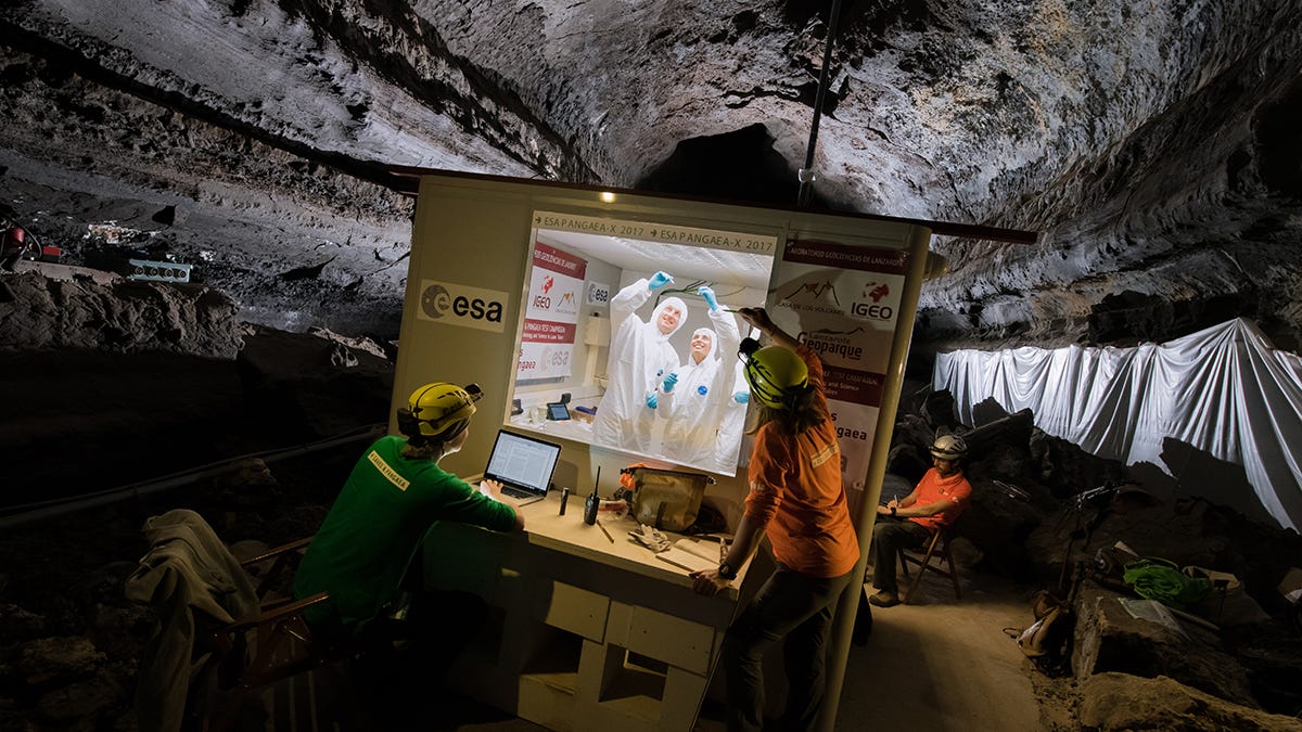 In what appears to be a cave, there is a clean room holding scientists in lab coats and hair nets. Outside the sort of box, there are a couple of scientists without lab coats on, exposed to the cave&apos;s environment.