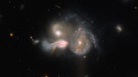 Three galaxies. They are close enough that they appear to be merging into one. Their shapes are distorted, with strands of gas and dust running between them. Each is emitting a lot of light. The background is dark, with a few smaller, dim and faint galaxies and a couple of stars.