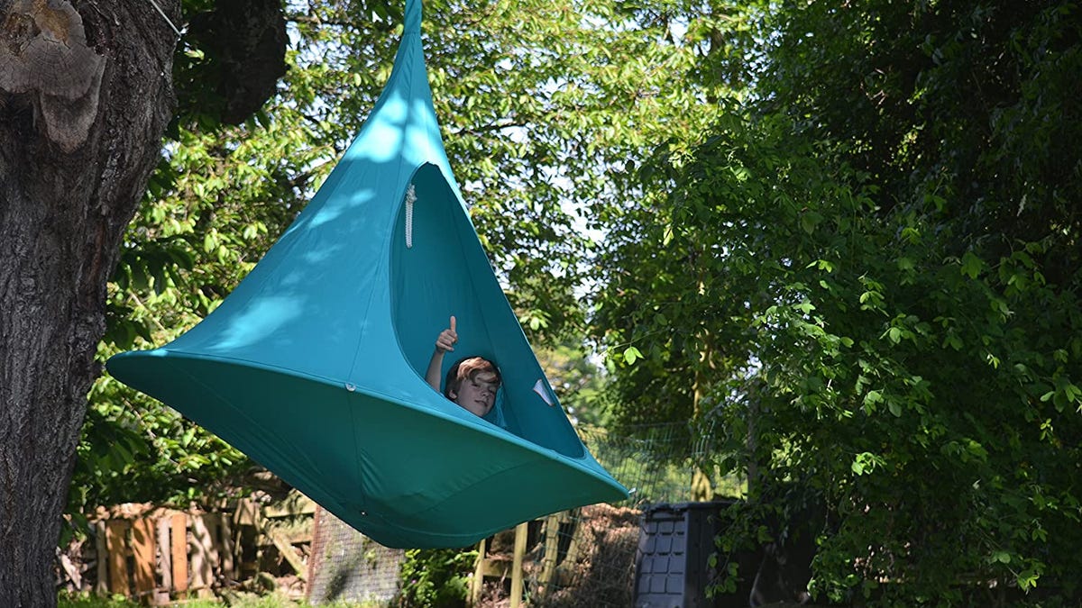A person sitting in a turquoise Vivere cacoon hammock hanging from tree.