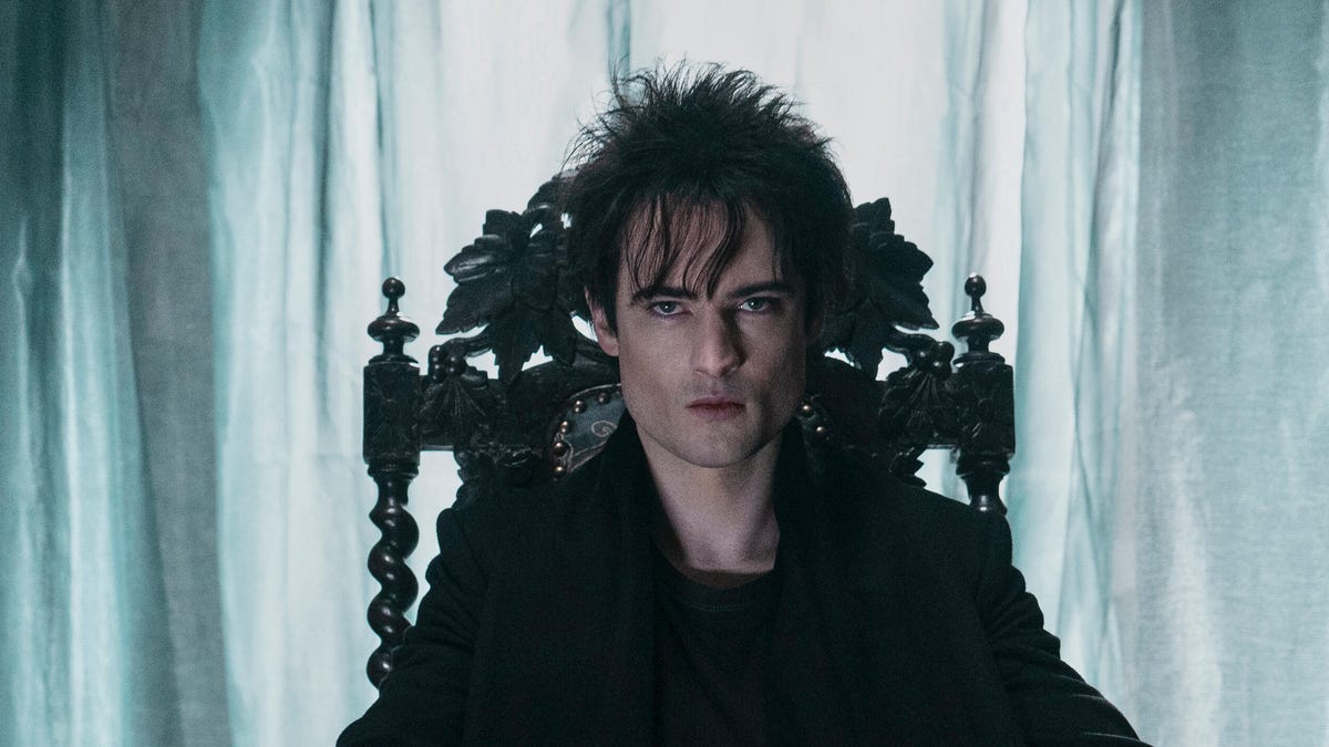 A man with great cheekbones and very black hair gazes down from a gothic throne.