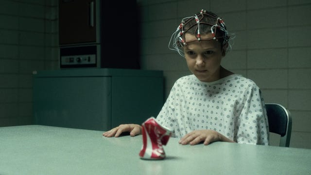 Stranger Things character Eleven stares intently at a crushed can of Coca-Cola while wearing sensors all over her head.