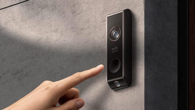 A hand points toward the Eufy dual-cam doorbell, which is installed on a concrete wall.