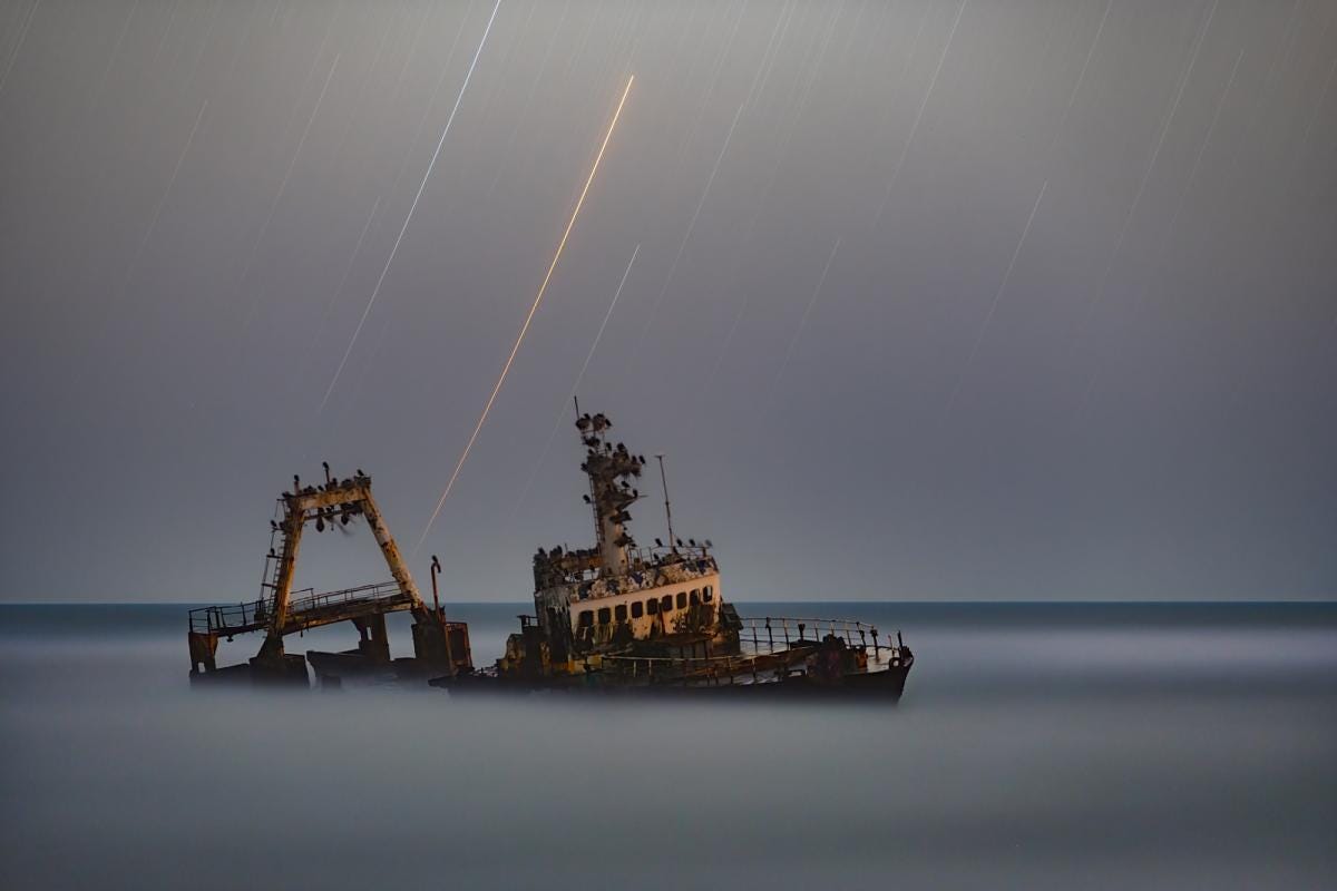 Soft gray water with a skeletonized shipwreck at an angle with bright star trails in the sky above.