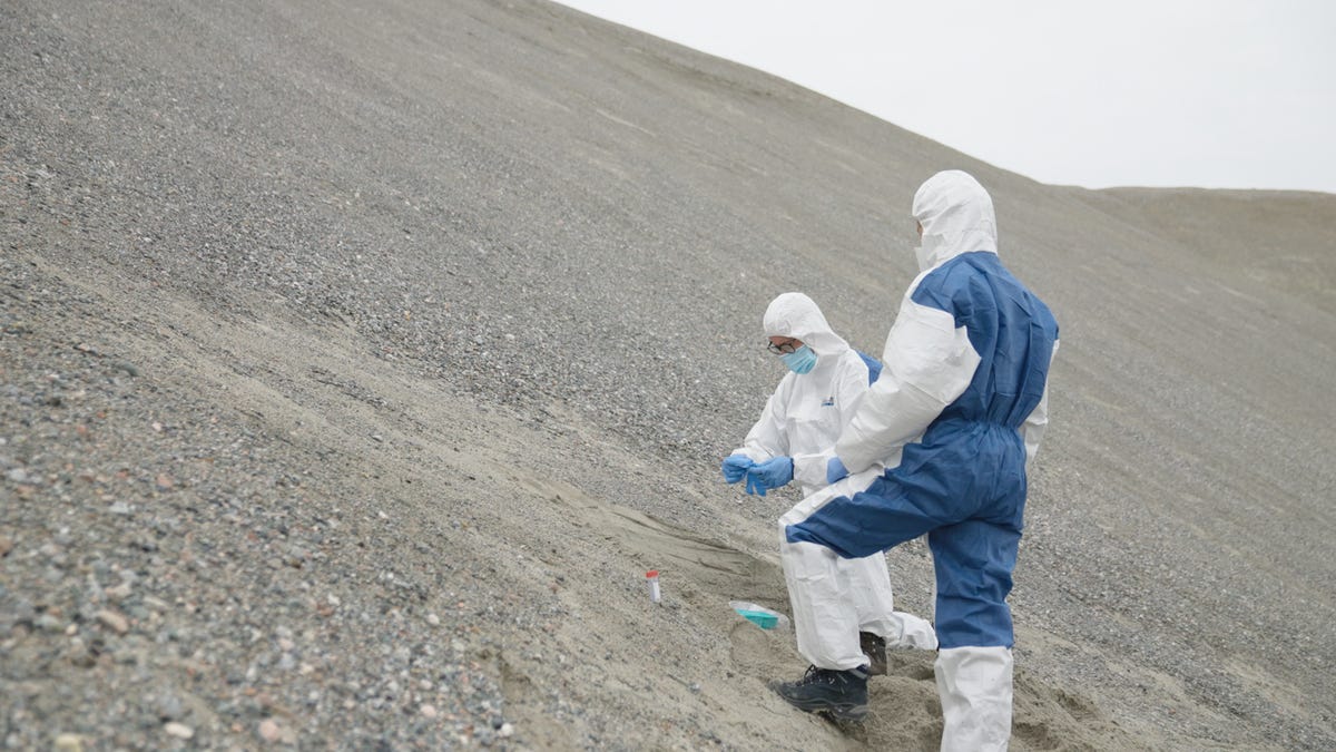 Two people stand on a dull gray slope wearing white protective gear that covers their entire body as they take soil samples.