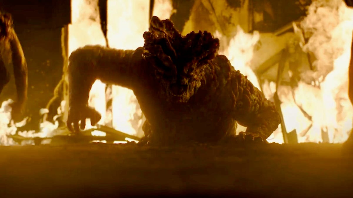 A scary, nasty Last of Us creature crawls up out of the ground and toward the camera as flames rage in the background.