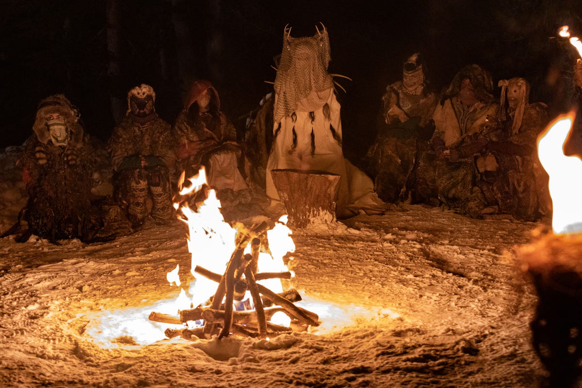 A group of girls sits around a bonfire wearing antlers and masks.