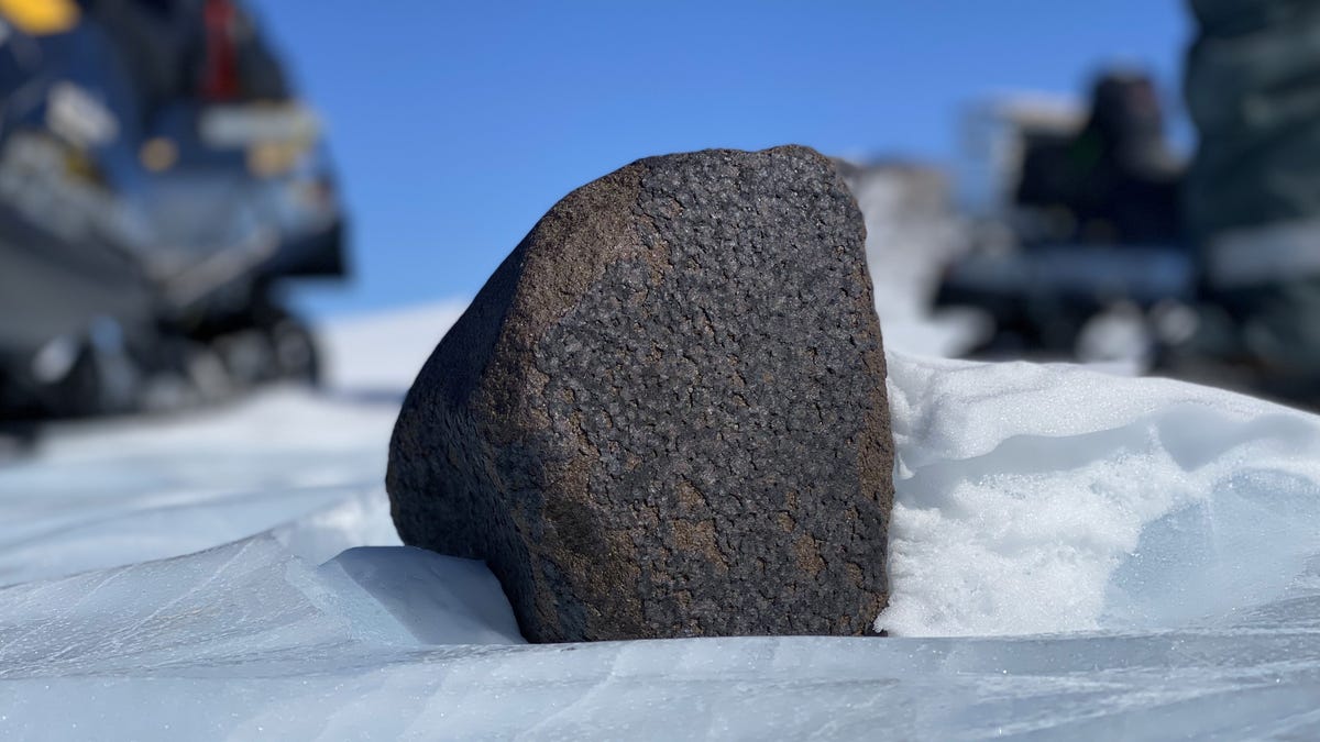 Large dark meteorite sits on white snow in Antarctica.