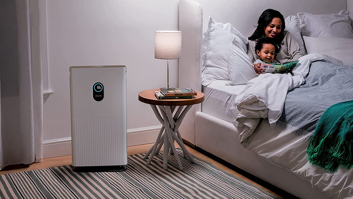 A woman reads a book to a little boy as they lie in bed and the Shark HE601 Air Purifier 6 rests nearby.
