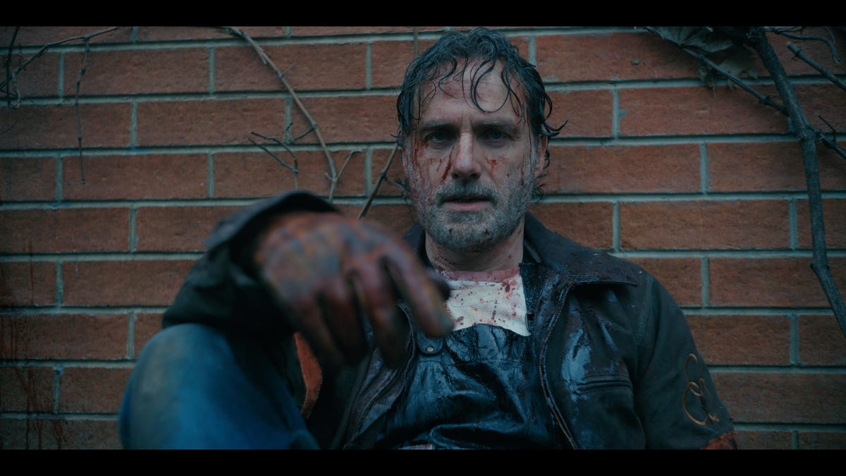 Andrew Lincoln como Rick Grimes sentado contra una pared de ladrillos con una mancha de sangre en la cara.
