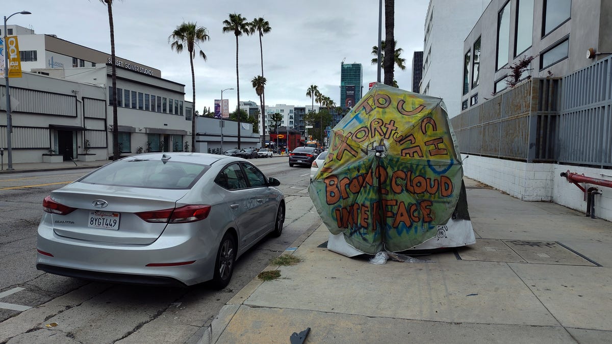 A colorful, decorated umbrella with vaguely apocalyptic text scrawled on it sits on a sidewalk under a gray Los Angeles sky.
