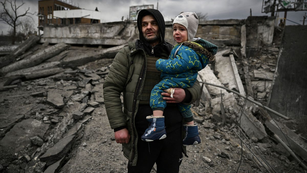 A man holding a child stands amid the rubble of a destroyed building. The man's fingers are bandaged, and the child, who wears a hood decorated like a teddy bear, is crying.