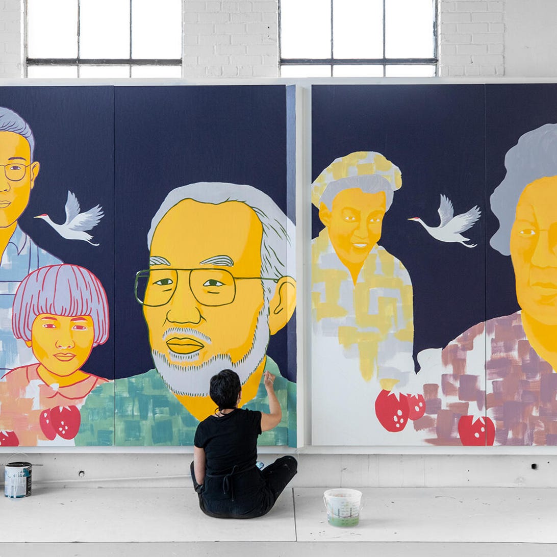 A woman sits cross-legged on the floor painting a wall-sized mural with faces of Japanese Canadian farmers.