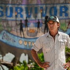 Jeff Probst stands with hands on hips in front of Survivor sign