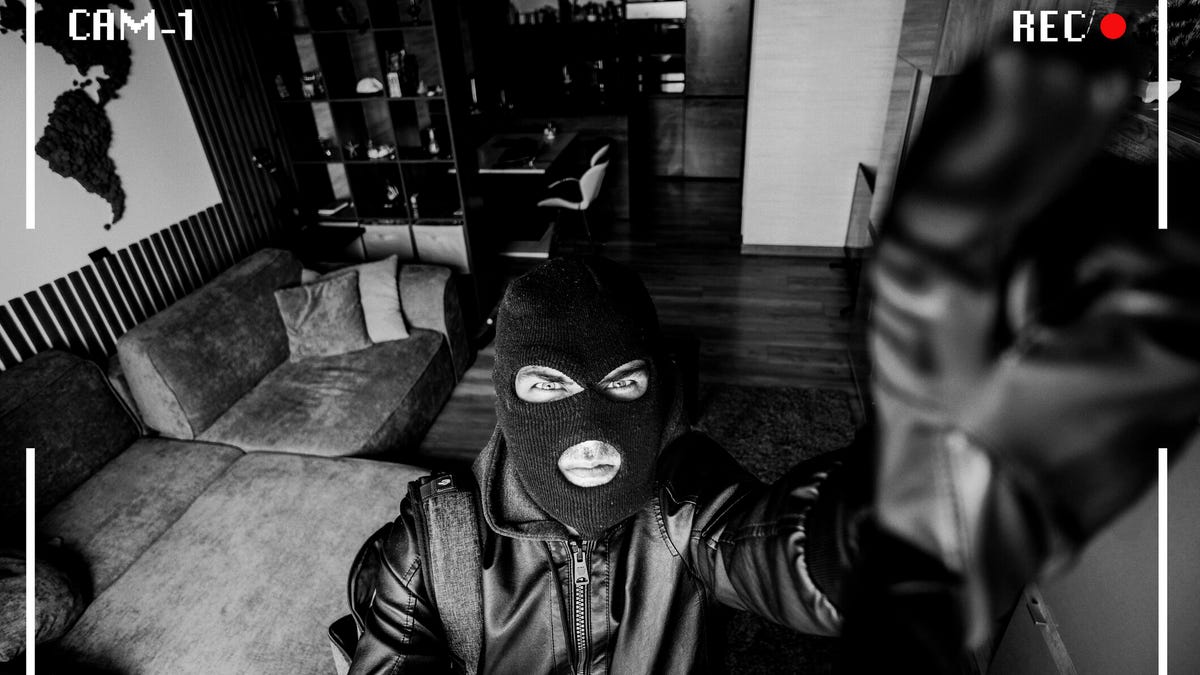 Mock picture of a security cam capturing a black-and-white image of a masked burglar fiddling with the lens.