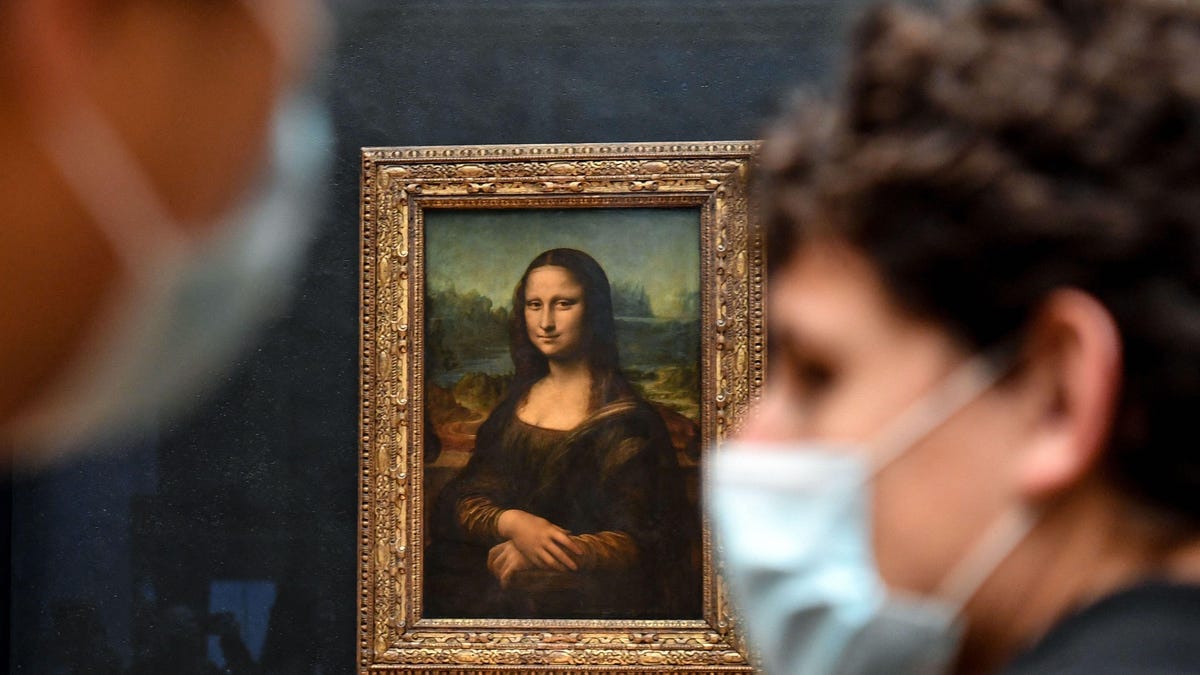 Visitors in medical masks walk past the painting The Mona Lisa by Leonardo Da Vinci hanging on a dark wall at The Louvre Museum in Paris.