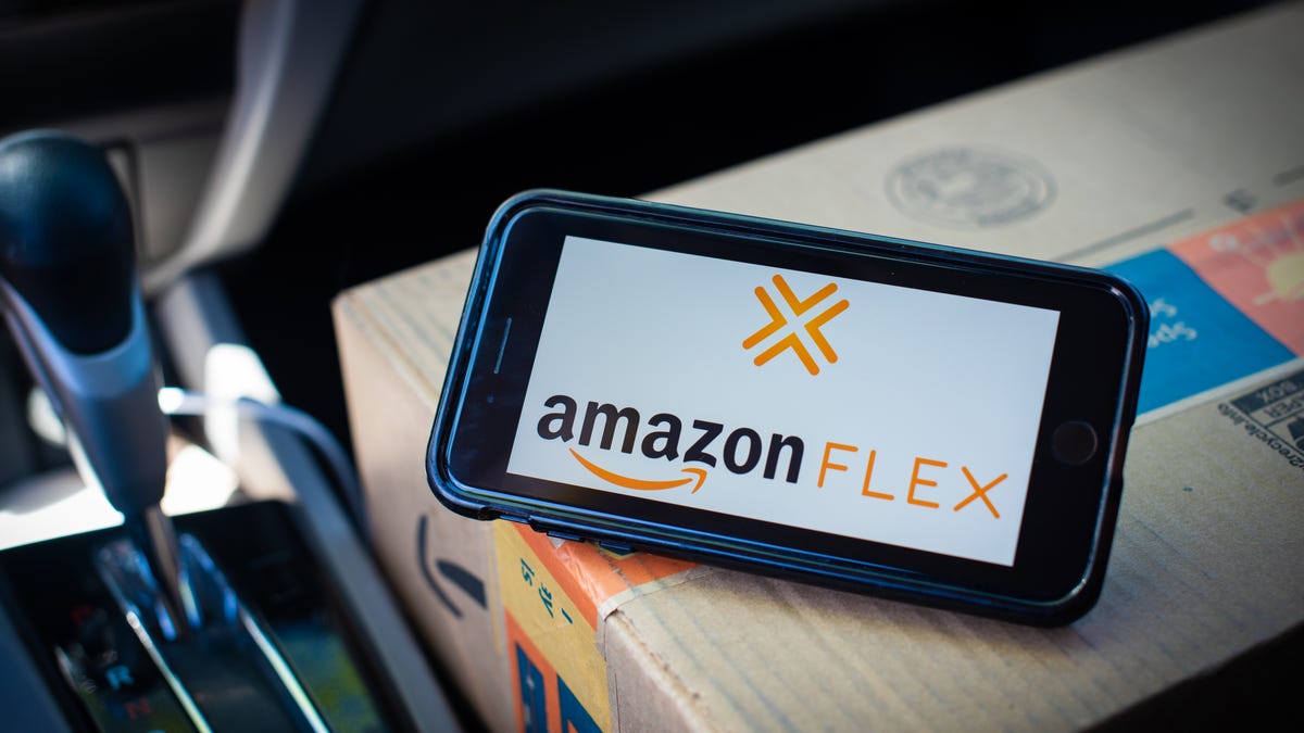The Amazon Flex logo is displayed on a a phone propped up on an Amazon box in the passenger seat of a car.