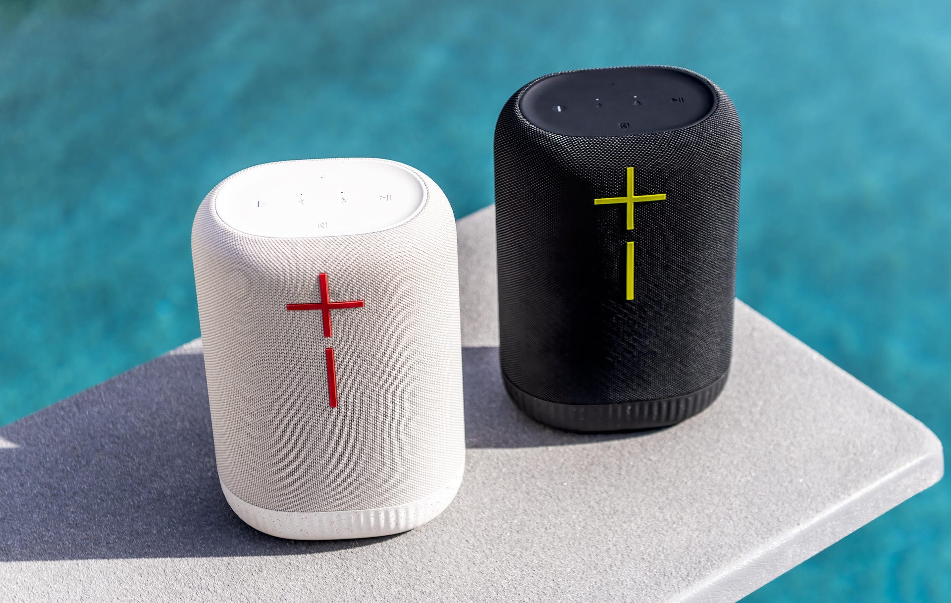 One black and one white UE Epicboom Bluetooth speaker, sitting on a diving board over a swimming pool
