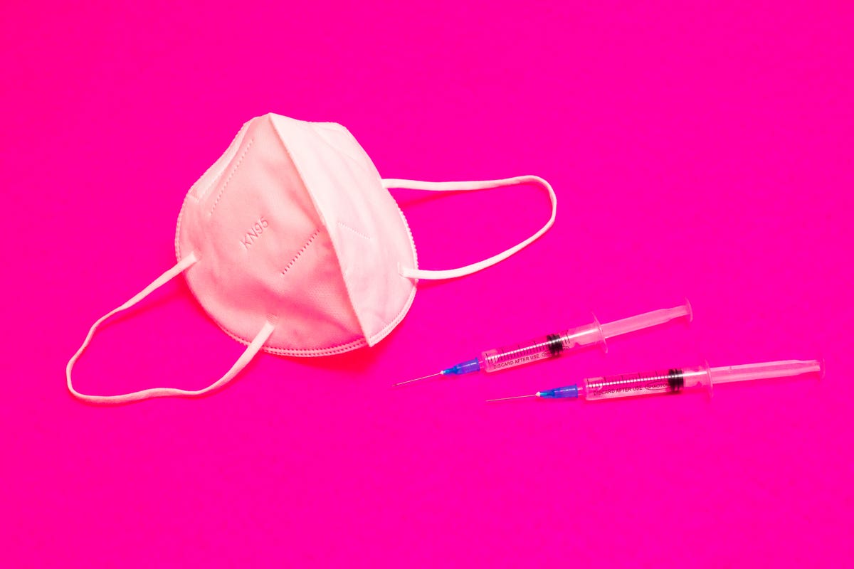 A face mask and two syringes on a neon-pink background.