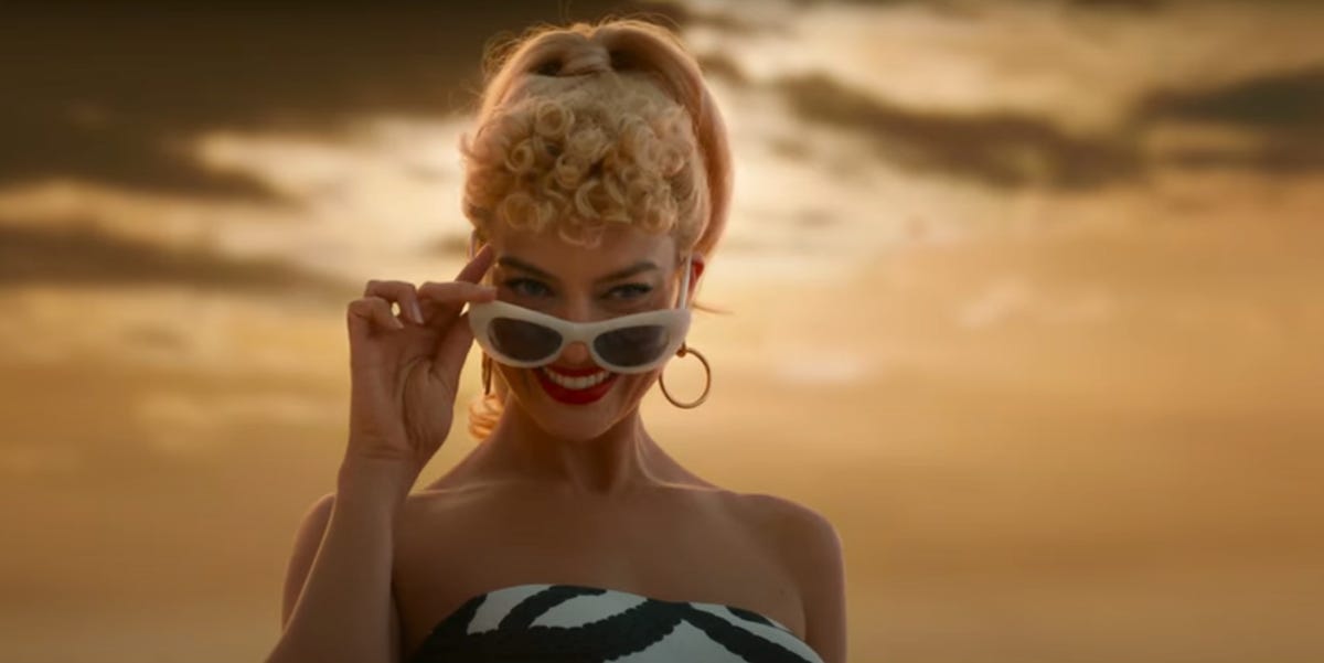 Margot Robbie smiles as she looks over her sunglasses with a cloudy sky in the background in the Barbie movie.