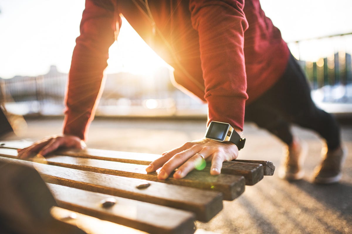 A man in a red shirt with a fitness watch on, doing push-ups on a park bench at sunset.