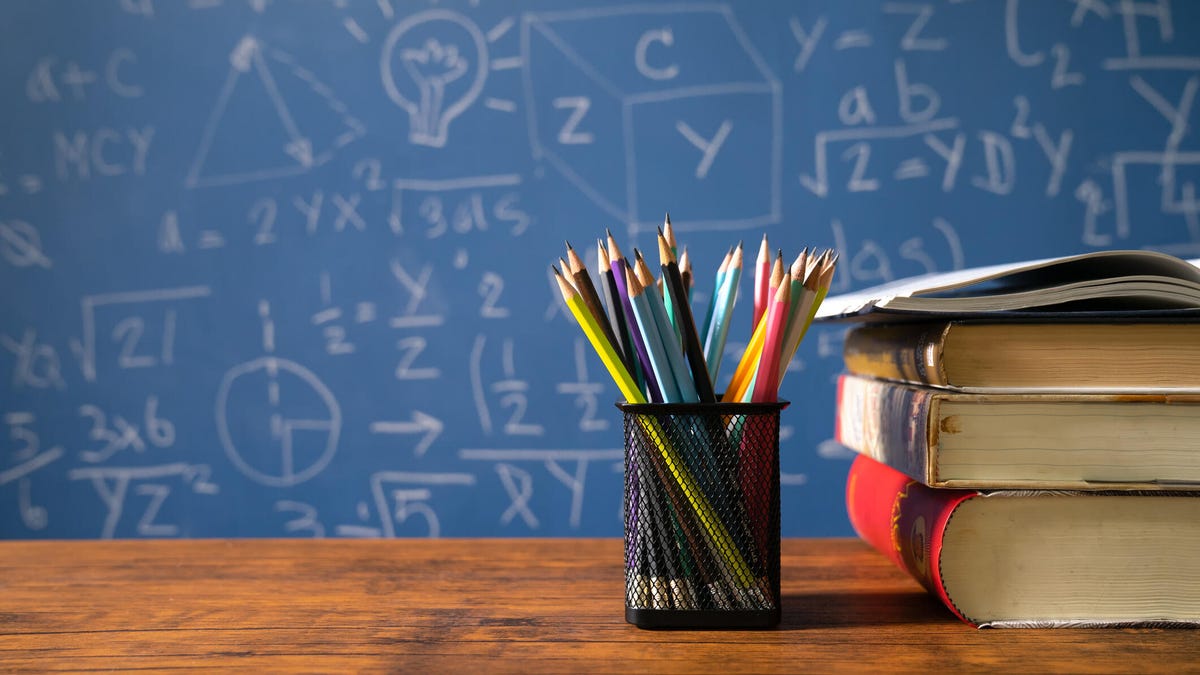A pencil cup and stack of books on a wooden desk with a blackboard in the background. 