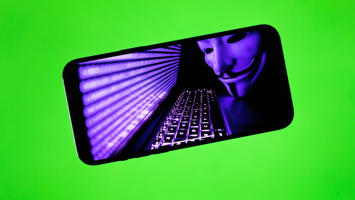 A Guy Fawkes mask next to an illuminated keyboard and a screen with blurred code