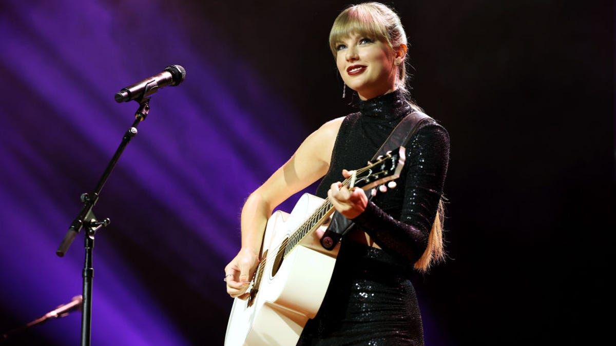Taylor Swift smiles onstage while playing a white acoustic guitar. She's wearing black, and the background is black but for several dramatic beams of violet light.