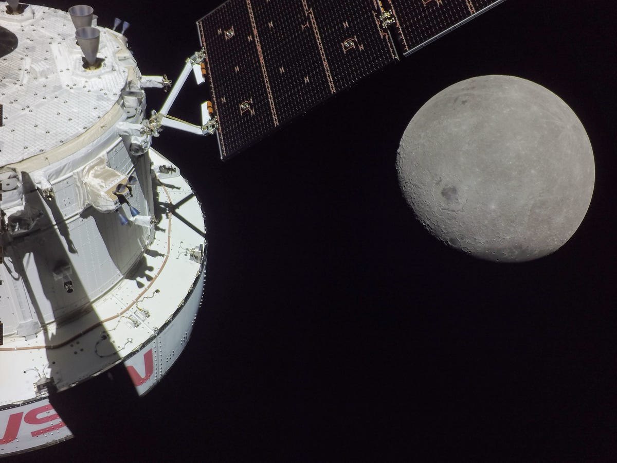 Космический корабль Орион выглядит беловатым с красным логотипом НАСА в виде червя и частью солнечной батареи, а луна вырисовывается частично освещенной вдалеке.