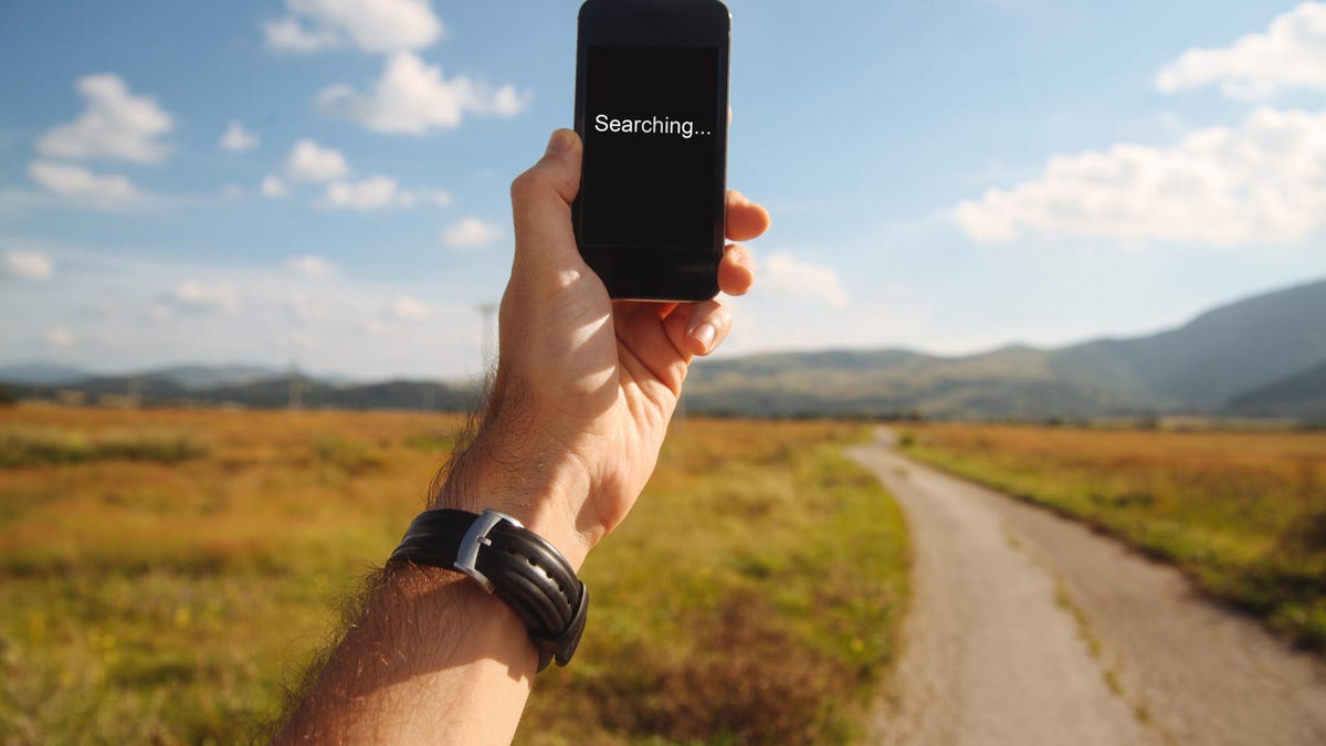 A hand holds up a phone with "Searching..." in text on its screen, with a rural road and wild background behind it.