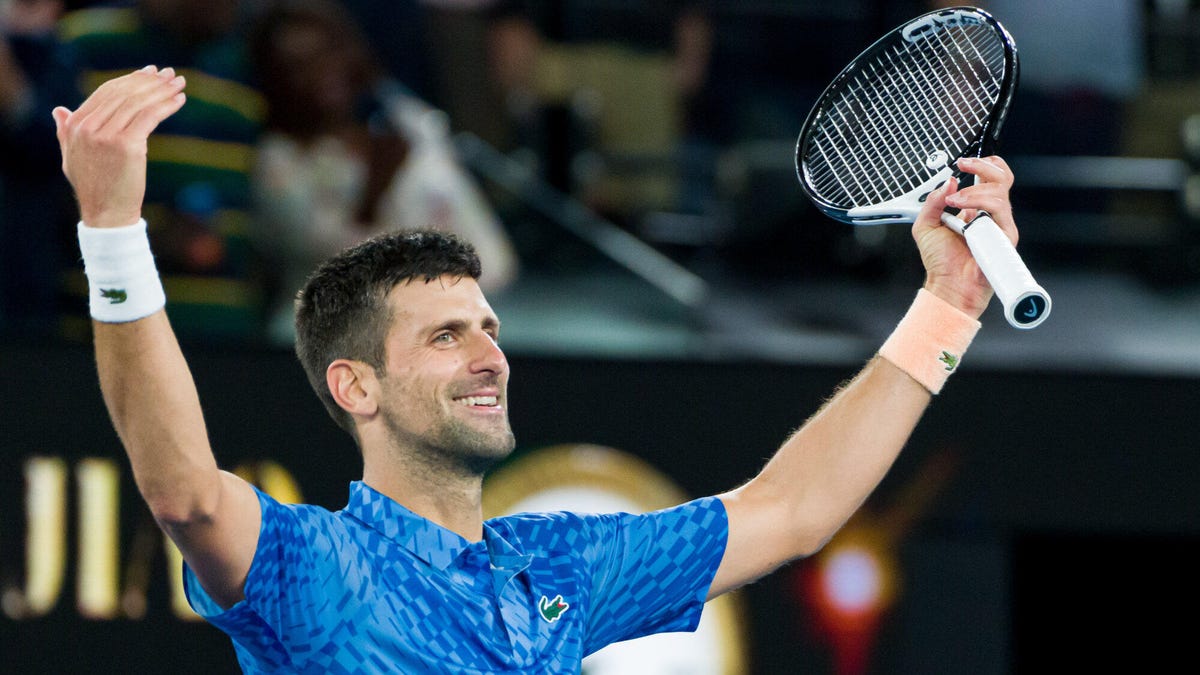 La estrella del tenis Novak Djokovic celebra con ambos brazos en alto y sostiene una raqueta en la mano izquierda.