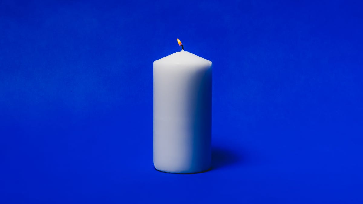 A lit white pillar candle in front of a plain blue background
