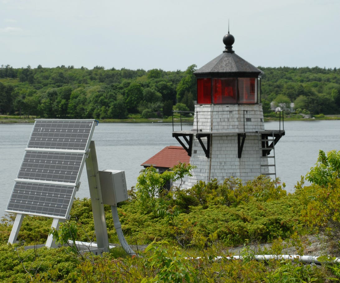 A ground mounted solar panel in front of a white lighthouse near water.