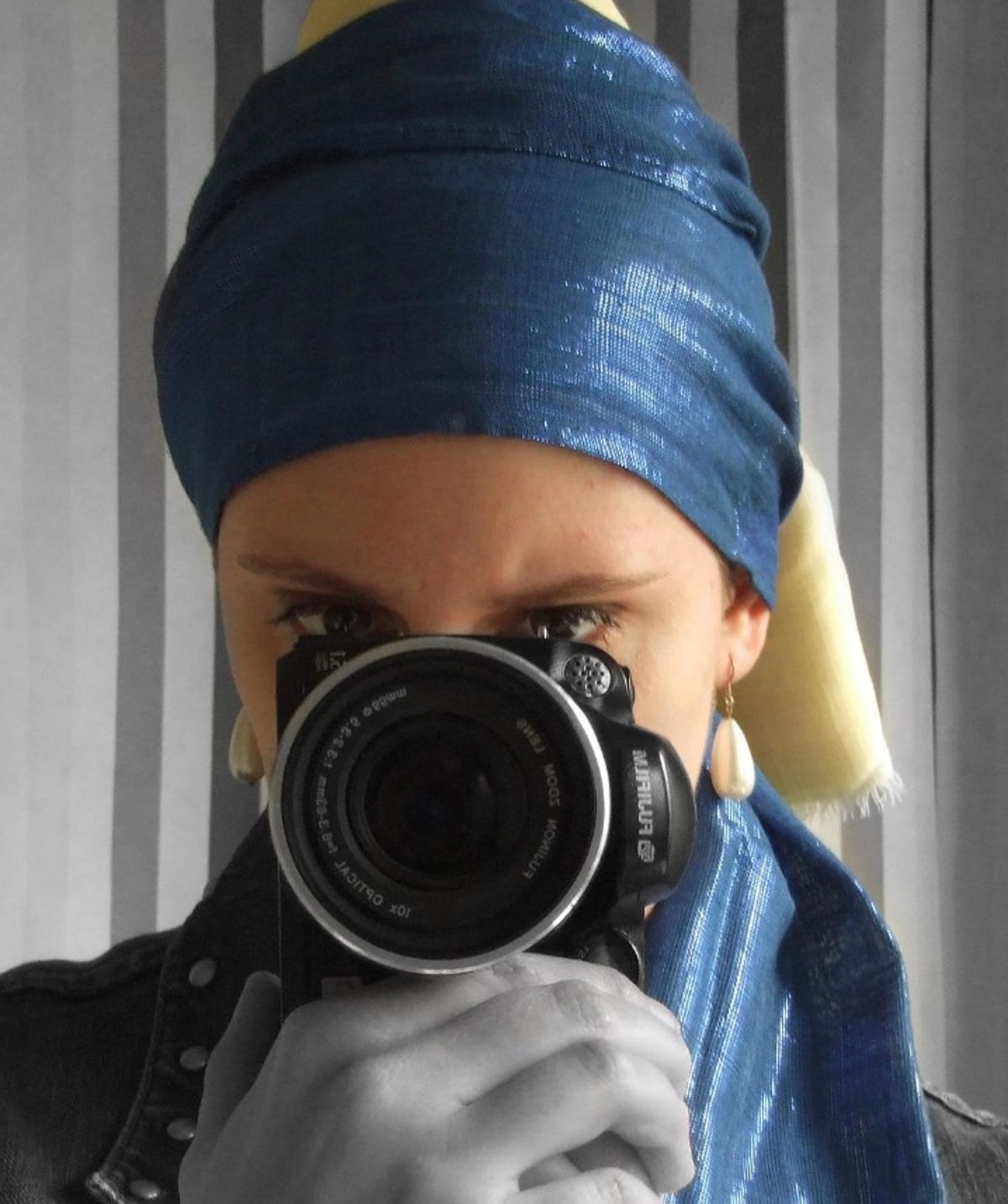 The woman wears a turban and two pearl earrings while holding a camera