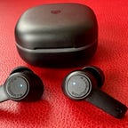 A pair of black SoundPEATS earbuds and charging case against a red background.