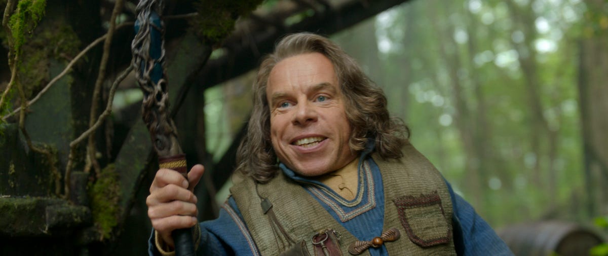 Actor Warwick Davis smiles and holds up a magic wand in a forest in fantasy TV series Willow.