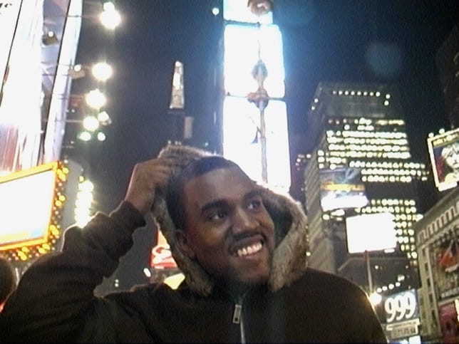 Kanye West smiles as he lowers the hood of his coat from his head. 