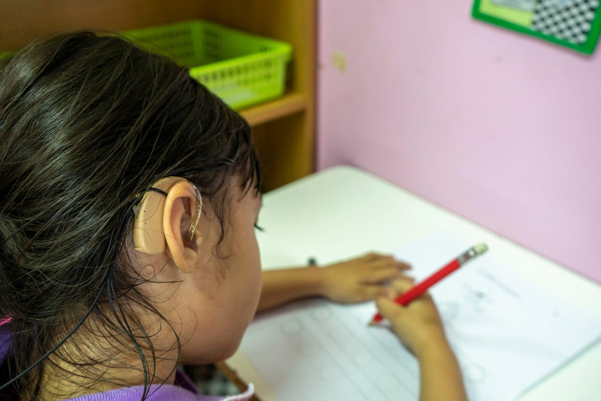 A child wearing a hearing aid writes with a pencil at her desk