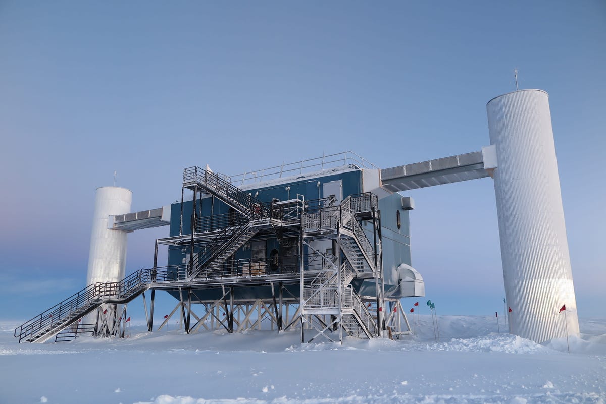 The IceCube observatory at the South Pole, surrounded by snow, with a central rectangular unit and two cylindrical towers on either side