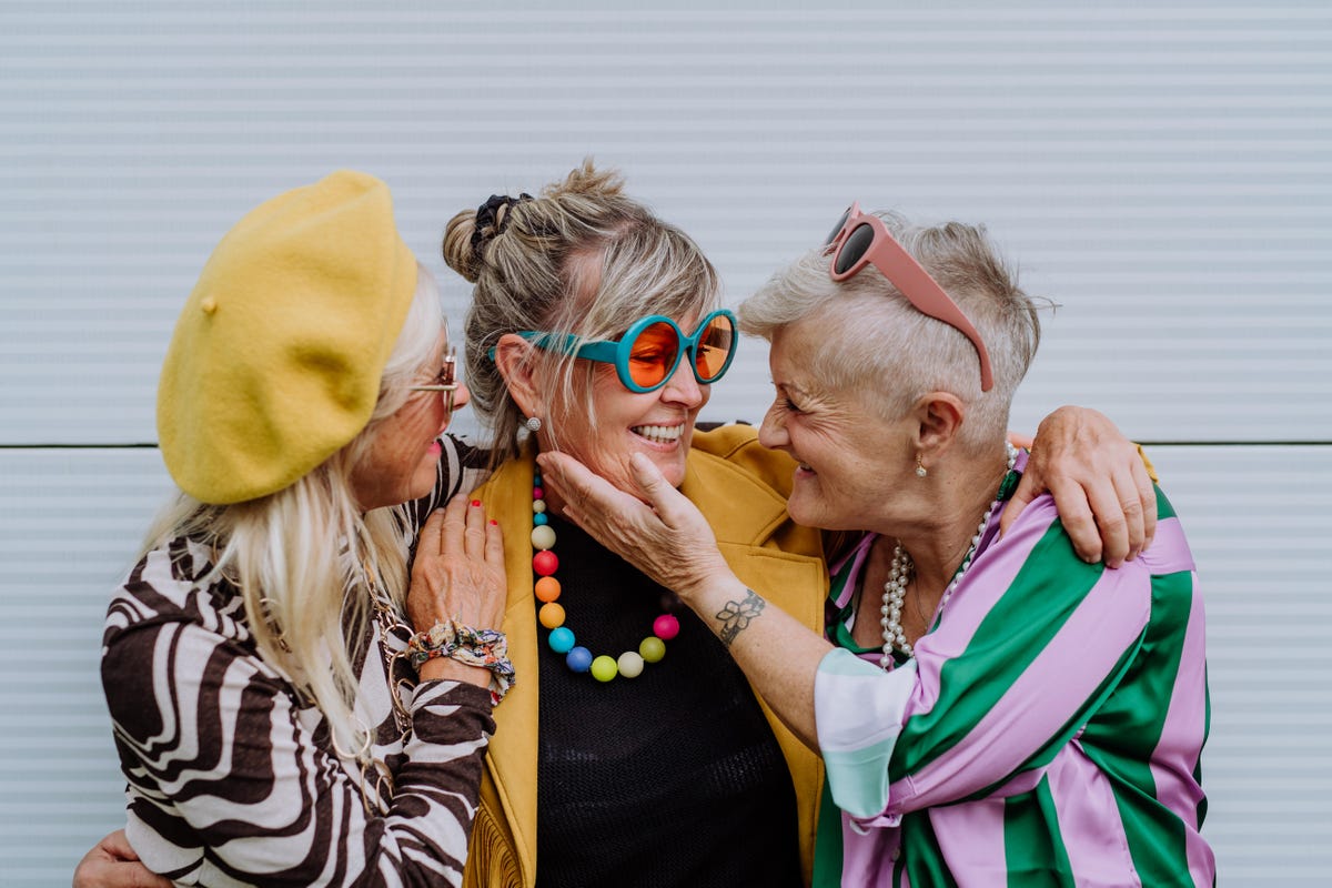 Three mature friends smile at each other wearing colorful glasses, hats and beads