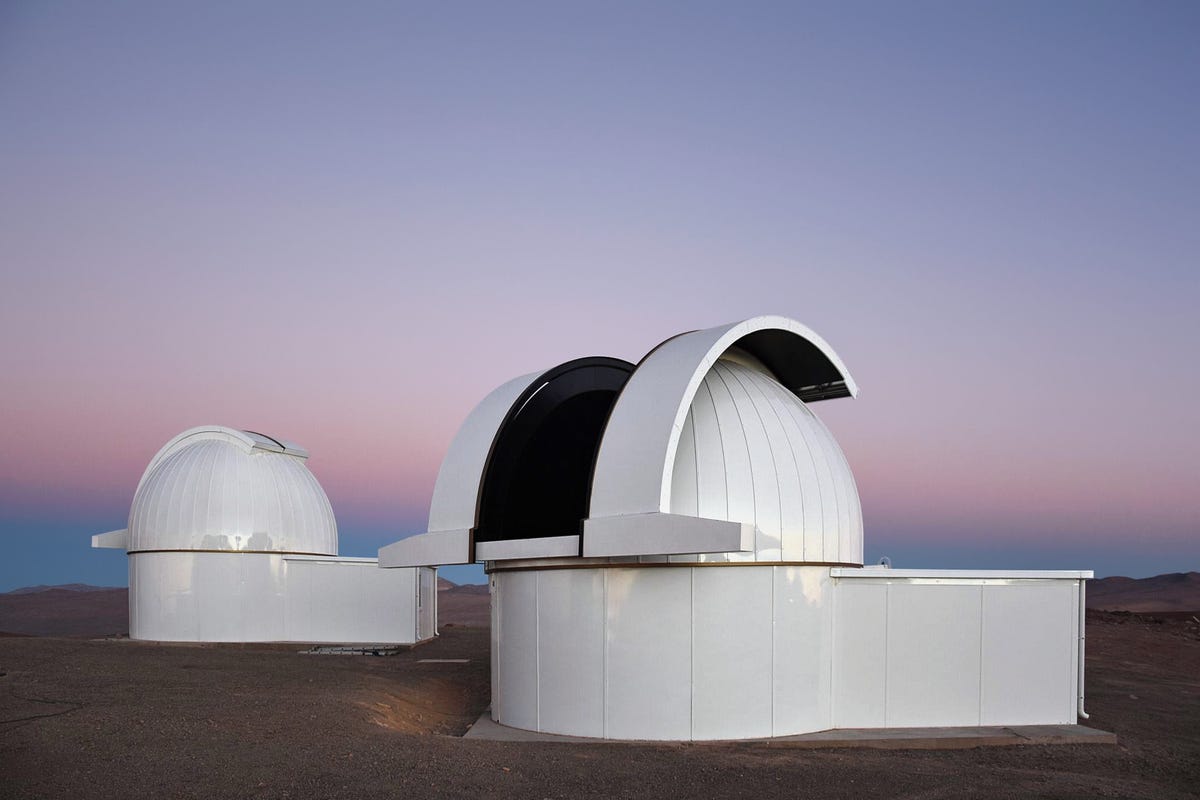 Two white domes are under the sky, on what looks like a rocky desert ground. These are some of the Speculoos telescopes.