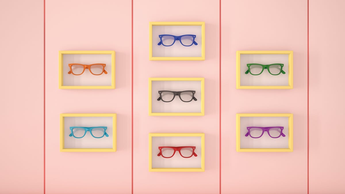 seven pairs of colorful eyeglasses sit in yellow rectangular frames on a pink wall